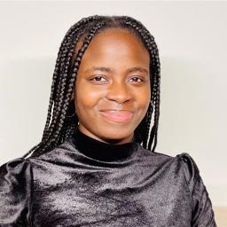 Vanessa, centered in the photo, captured shoulder up, smiling. Vanessa has braids and is wearing a black top. She is infront of a white background.